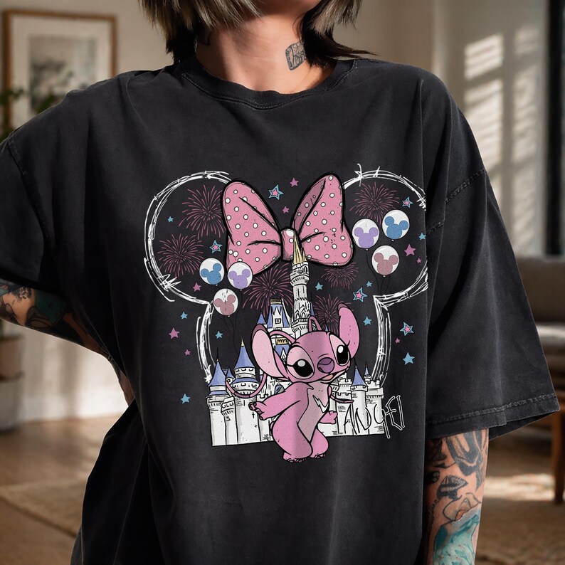 Puede incluir: Una camiseta de manga corta en color negro carb&oacute;n con un estampado gr&aacute;fico de un personaje alien&iacute;gena rosa frente al contorno de un castillo blanco. El dise&ntilde;o incluye un gran lazo rosa con lunares, fuegos artificiales y globos circulares.