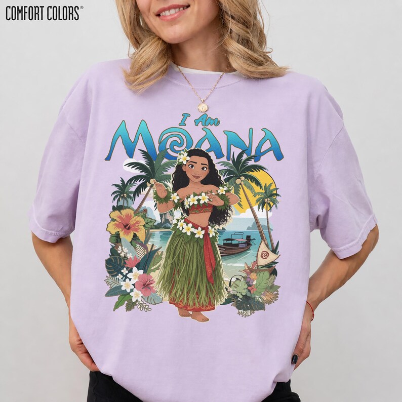 May include: A light purple t-shirt with the words "I Am Moana" in blue and green. The graphic features a cartoon of Moana in a grass skirt and flower lei, with palm trees and a boat in the background. The brand "Comfort Colors" is printed at the top.