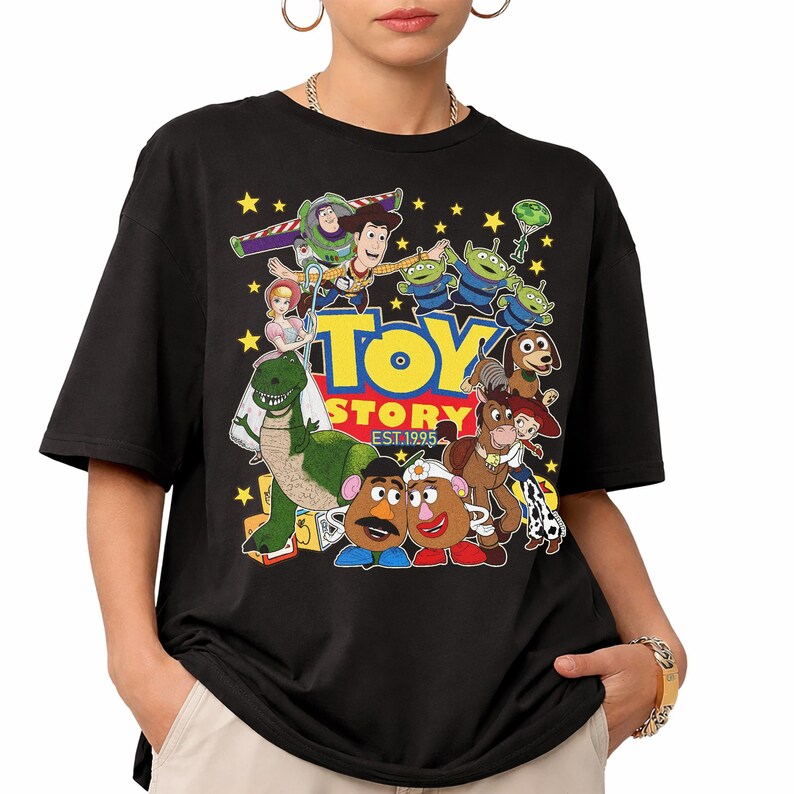 May include: Black t-shirt featuring a colorful graphic of characters from the animated film "Toy Story." The design includes Buzz Lightyear, Woody, and other characters, with the text "TOY STORY EST.1995" in yellow and red.