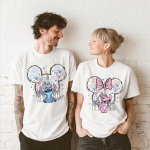 Puede incluir: Dos camisetas blancas de manga corta con estampados gr&aacute;ficos de personajes de Disney. La camiseta de la izquierda muestra al personaje Stitch azul con un castillo y fuegos artificiales dentro de una silueta de Mickey Mouse. La derecha muestra a Angel.