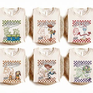 May include: Six off-white t-shirts featuring Toy Story characters: Aliens, Woody, Forky, Slinky, Jessie, and Buzz. Each shirt has a character graphic, the character's name, and the year 1995 printed on it. The shirts are displayed on woven mats.
