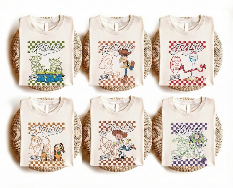 May include: Six off-white t-shirts featuring Toy Story characters: Aliens, Woody, Forky, Slinky, Jessie, and Buzz. Each shirt has a character graphic, the character's name, and the year 1995 printed on it. The shirts are displayed on woven mats.