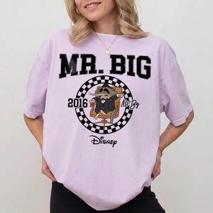 May include: A light purple short-sleeve t-shirt featuring a graphic of a cartoon shrew in a suit sitting in a chair. The design includes a black and white checkered circle, the text MR. BIG, the year 2016, the name Mr. Big, and the Disney logo.