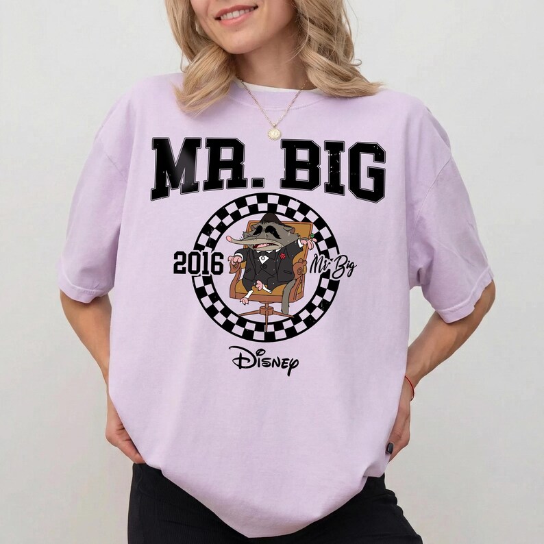 May include: A light purple short-sleeve t-shirt featuring a graphic of a cartoon shrew in a suit sitting in a chair. The design includes a black and white checkered circle, the text MR. BIG, the year 2016, the name Mr. Big, and the Disney logo.