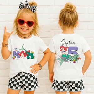 May include: Two white t-shirts with a mermaid theme. One shirt has the word "MOM" with a mermaid graphic. The other shirt has the name "Sophia" and the number "5" with a mermaid graphic. The girls are wearing black and white checkered shorts.