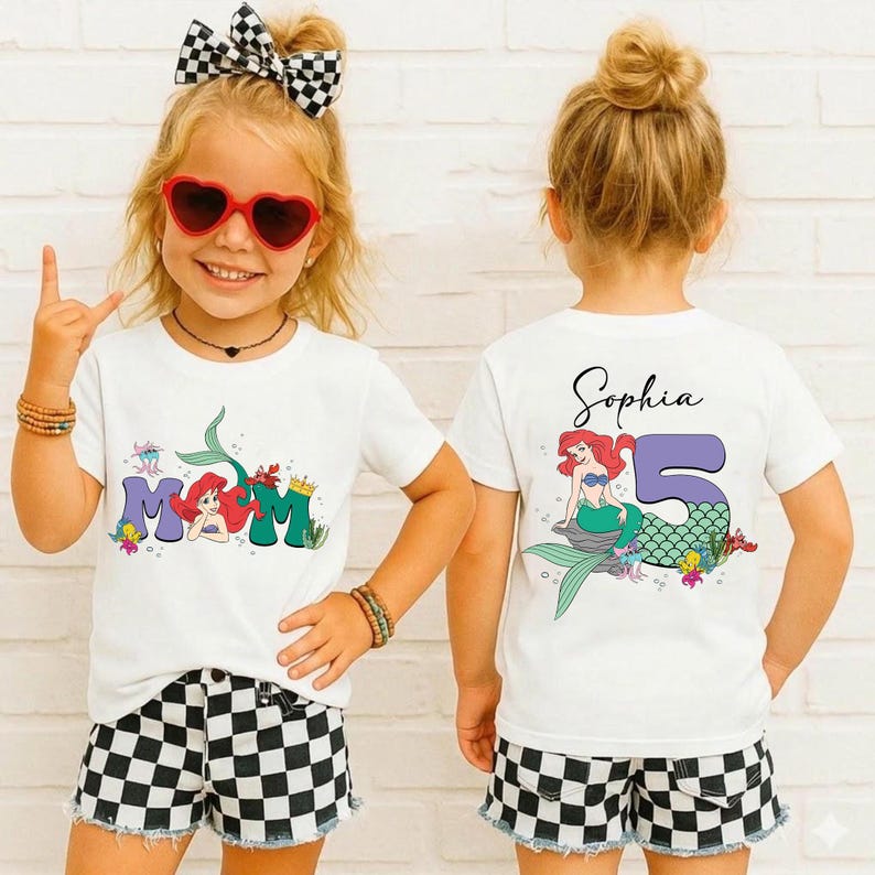 May include: Two white t-shirts with a mermaid theme. One shirt has the word "MOM" with a mermaid graphic. The other shirt has the name "Sophia" and the number "5" with a mermaid graphic. The girls are wearing black and white checkered shorts.