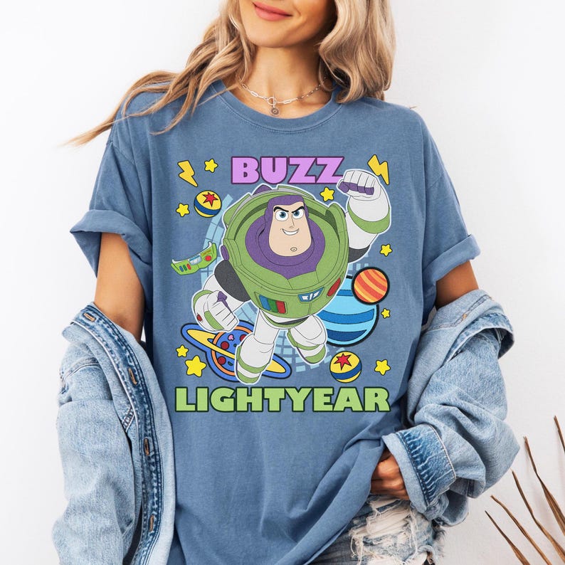 May include: Blue t-shirt featuring a graphic of Buzz Lightyear from Toy Story. The design includes the text "BUZZ LIGHTYEAR" in purple and green, with cartoon planets and stars. The shirt has rolled-up sleeves.