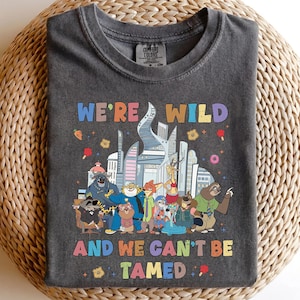 K&ouml;nnte beinhalten: Ein anthrazitfarbenes T-Shirt mit Rundhalsausschnitt und einer bunten Grafik verschiedener animierter Tierfiguren vor einer Stadtsilhouette. Der Text "WE'RE WILD" steht oben und "AND WE CAN'T BE TAMED" unten in mehrfarbigen Buchstaben.