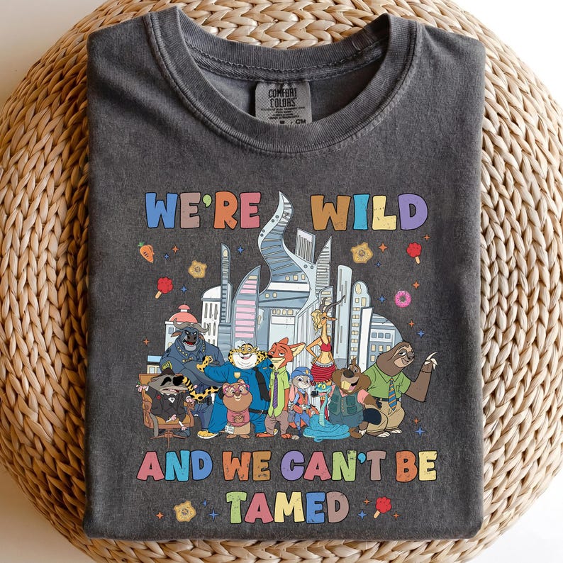 K&ouml;nnte beinhalten: Ein anthrazitfarbenes T-Shirt mit Rundhalsausschnitt und einer bunten Grafik verschiedener animierter Tierfiguren vor einer Stadtsilhouette. Der Text "WE'RE WILD" steht oben und "AND WE CAN'T BE TAMED" unten in mehrfarbigen Buchstaben.