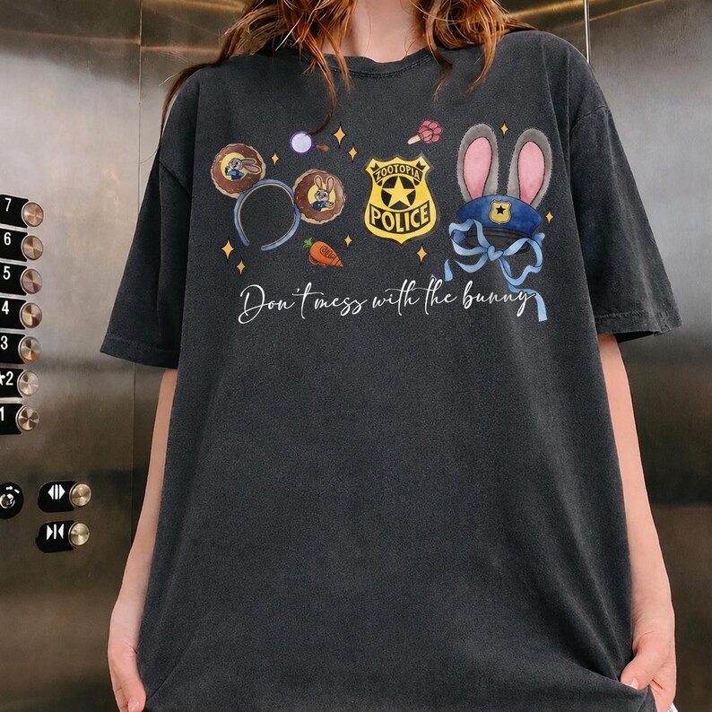 May include: A charcoal gray t-shirt featuring a graphic print with Zootopia-themed elements including mouse ears, a police badge, a carrot, and a blue hat with rabbit ears. The text "Don't mess with the bunny" is printed in white script.