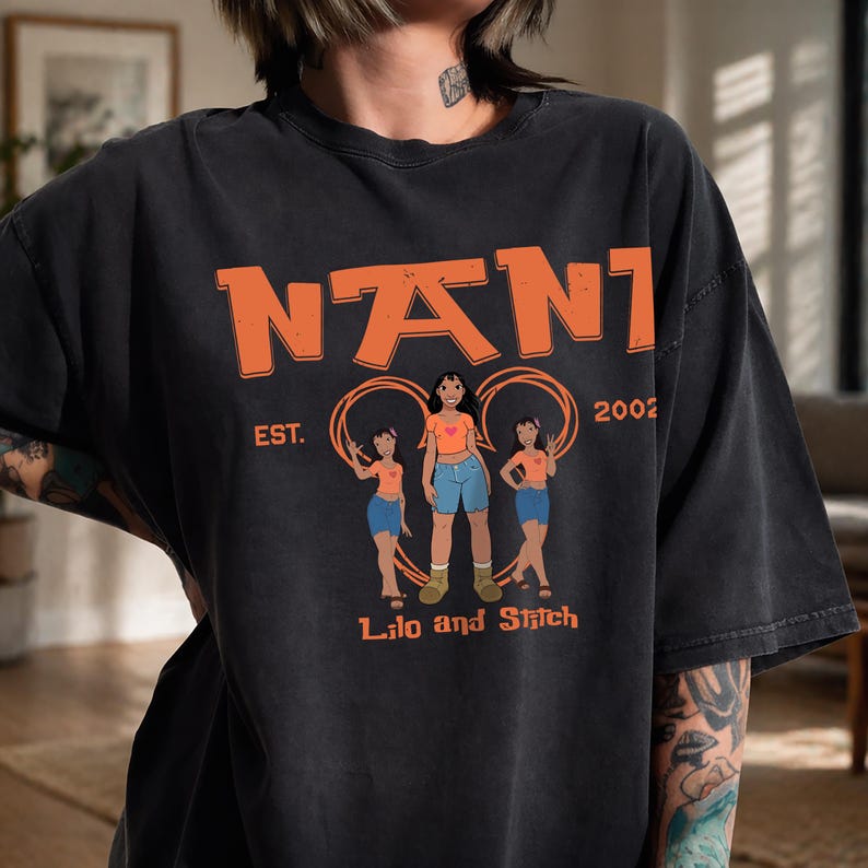 Puede incluir: Una camiseta de manga corta gris carb&oacute;n con un estampado gr&aacute;fico que incluye el texto naranja NANI en la parte superior, EST. 2002 a los lados y Lilo and Stitch en la parte inferior. El dise&ntilde;o incluye tres ilustraciones de Nani con camisa naranja y shorts azules.