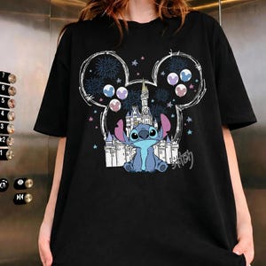 Puede incluir: Una camiseta negra de manga corta con un estampado gr&aacute;fico del personaje Stitch sentado frente al contorno de un castillo blanco. El dise&ntilde;o est&aacute; enmarcado por una gran silueta de orejas de Mickey Mouse llena de fuegos artificiales, estrellas y globos.