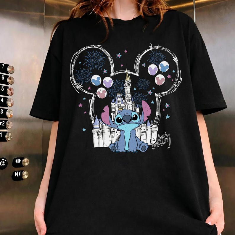 Puede incluir: Una camiseta negra de manga corta con un estampado gr&aacute;fico del personaje Stitch sentado frente al contorno de un castillo blanco. El dise&ntilde;o est&aacute; enmarcado por una gran silueta de orejas de Mickey Mouse llena de fuegos artificiales, estrellas y globos.