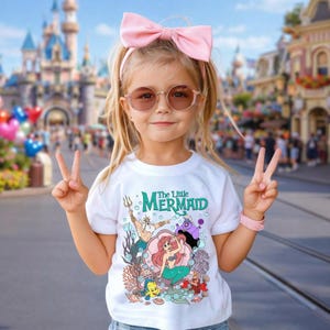 Puede incluir: Una ni&ntilde;a peque&ntilde;a con una camiseta blanca con un gr&aacute;fico colorido de personajes de "La Sirenita". La camiseta presenta a Ariel, el Rey Trit&oacute;n, &Uacute;rsula y otros personajes. Tambi&eacute;n lleva un lazo rosa, gafas de sol y una pulsera rosa.