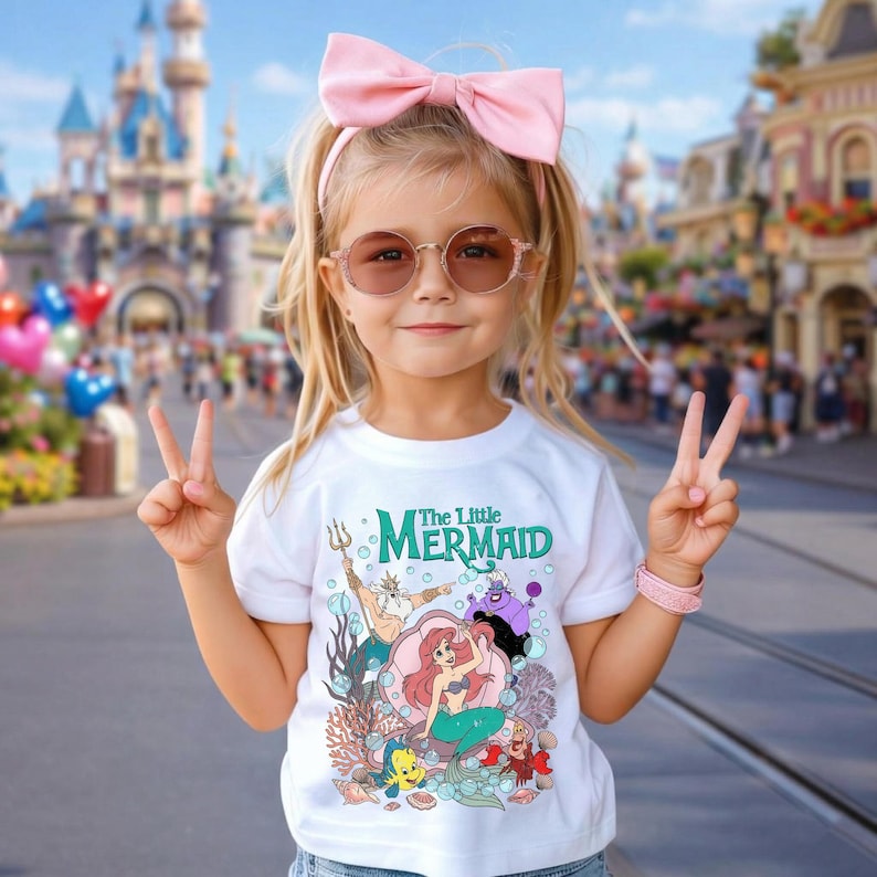 Puede incluir: Una ni&ntilde;a peque&ntilde;a con una camiseta blanca con un gr&aacute;fico colorido de personajes de "La Sirenita". La camiseta presenta a Ariel, el Rey Trit&oacute;n, &Uacute;rsula y otros personajes. Tambi&eacute;n lleva un lazo rosa, gafas de sol y una pulsera rosa.