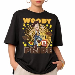 May include: Black t-shirt featuring a graphic of Woody from Toy Story. The design includes the word "WOODY" above the character and "PRIDE" below, with stars and lightning bolts. Woody is depicted in his cowboy attire.