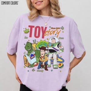 May include: A light purple Comfort Colors t-shirt featuring a graphic of characters from the movie Toy Story. The graphic includes the text "TOY STORY" and the characters Rex, Woody, and Buzz Lightyear.