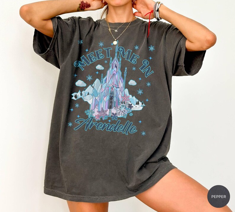 May include: Dark gray t-shirt with a graphic featuring a blue and purple castle, mountains, and the text "Meet Me In Arendelle." The design includes snowflakes and floral accents. The shirt has short sleeves and a relaxed fit.