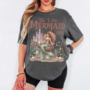 May include: Dark gray t-shirt featuring a graphic of Ariel from The Little Mermaid. The design includes a castle, seashell throne, and the text "The Little Mermaid: A Vintage Sea Tale" in brown lettering. The shirt has short sleeves.