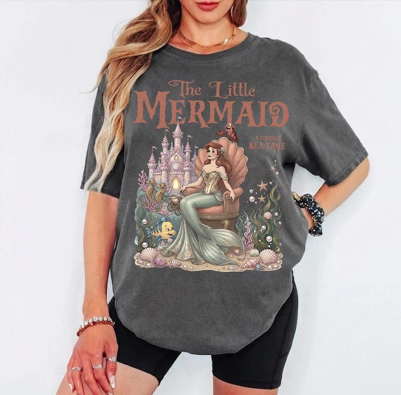 May include: Dark gray t-shirt featuring a graphic of Ariel from The Little Mermaid. The design includes a castle, seashell throne, and the text "The Little Mermaid: A Vintage Sea Tale" in brown lettering. The shirt has short sleeves.