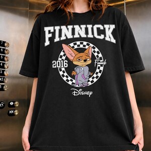May include: A black short-sleeved t-shirt featuring a graphic of the character Finnick from Disney. The design includes the text FINNICK, 2016, and Disney, centred within a black and white chequered circle. The character wears a grey elephant costume.