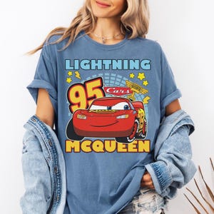 May include: Blue t-shirt featuring a graphic of Lightning McQueen from the Cars movie. The design includes the number 95, stars, and the text "Lightning McQueen" and "Cars". The shirt has rolled-up sleeves.