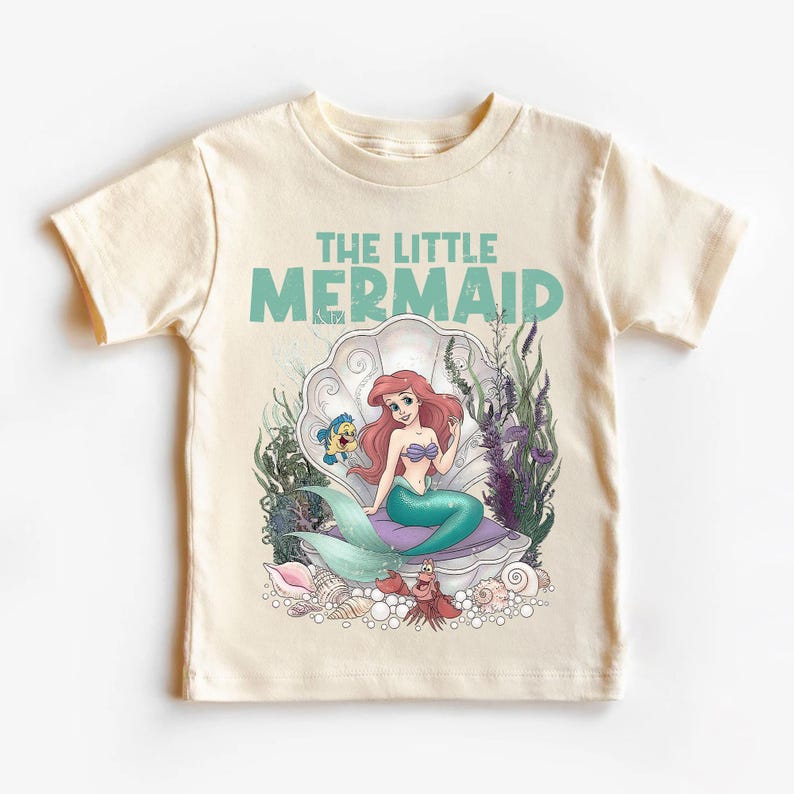 May include: A cream-colored t-shirt featuring a graphic of Ariel from The Little Mermaid. The design includes Ariel sitting in a seashell, with Flounder and Sebastian. The words "THE LITTLE MERMAID" are printed above the image in teal.