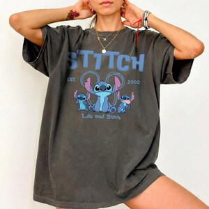 Puede incluir: Una camiseta gris carb&oacute;n de corte oversize con un estampado gr&aacute;fico en azul claro del personaje Stitch de Lilo y Stitch. El texto dice STITCH, EST. 2002 y Lilo and Stitch. El dise&ntilde;o incluye tres figuras azules de Stitch con orejas rosas.