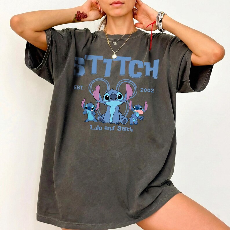Puede incluir: Una camiseta gris carb&oacute;n de corte oversize con un estampado gr&aacute;fico en azul claro del personaje Stitch de Lilo y Stitch. El texto dice STITCH, EST. 2002 y Lilo and Stitch. El dise&ntilde;o incluye tres figuras azules de Stitch con orejas rosas.