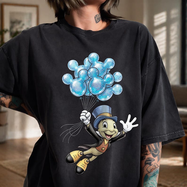 May include: Black t-shirt featuring a cartoon illustration of Jiminy Cricket holding a bunch of blue Mickey Mouse-shaped balloons. The balloons have a glittery effect. Jiminy Cricket is wearing a top hat and a suit.
