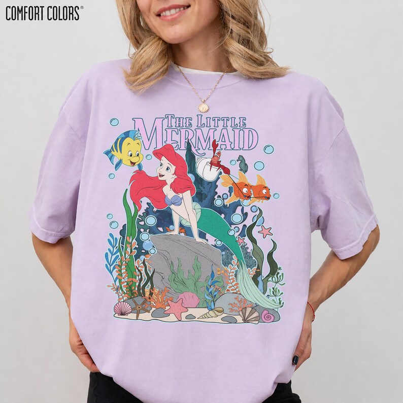 May include: A light purple t-shirt featuring a graphic of Ariel from The Little Mermaid. The design includes Ariel, Flounder, Sebastian, and other sea creatures. The words "The Little Mermaid" are printed above the characters in a purple font.