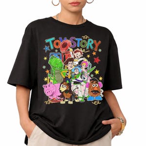 Może przedstawiać: Czarny t-shirt z kolorową grafiką postaci z filmu animowanego "Toy Story". Projekt zawiera Chudego, Buzz'a Astrala, Rexa i inne postacie, ze słowami "Toy Story" w zabawnej czcionce. Koszulka ma kr&oacute;tkie rękawy.