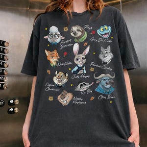May include: A charcoal gray t-shirt featuring a graphic print of various animated animal characters from Zootopia, including Judy Hopps, Nick Wilde, and Chief Bogo, with their names printed in white script alongside small yellow stars and orange carrots.