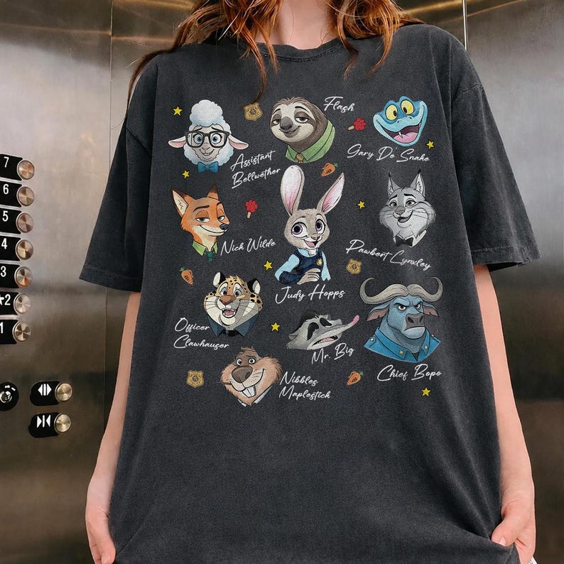 May include: A charcoal gray t-shirt featuring a graphic print of various animated animal characters from Zootopia, including Judy Hopps, Nick Wilde, and Chief Bogo, with their names printed in white script alongside small yellow stars and orange carrots.