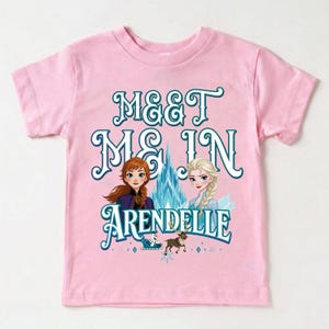 May include: Pink short-sleeved t-shirt featuring the text "MEET ME IN ARENDELLE" in teal script. The design includes illustrations of two animated characters, a castle, and a reindeer.