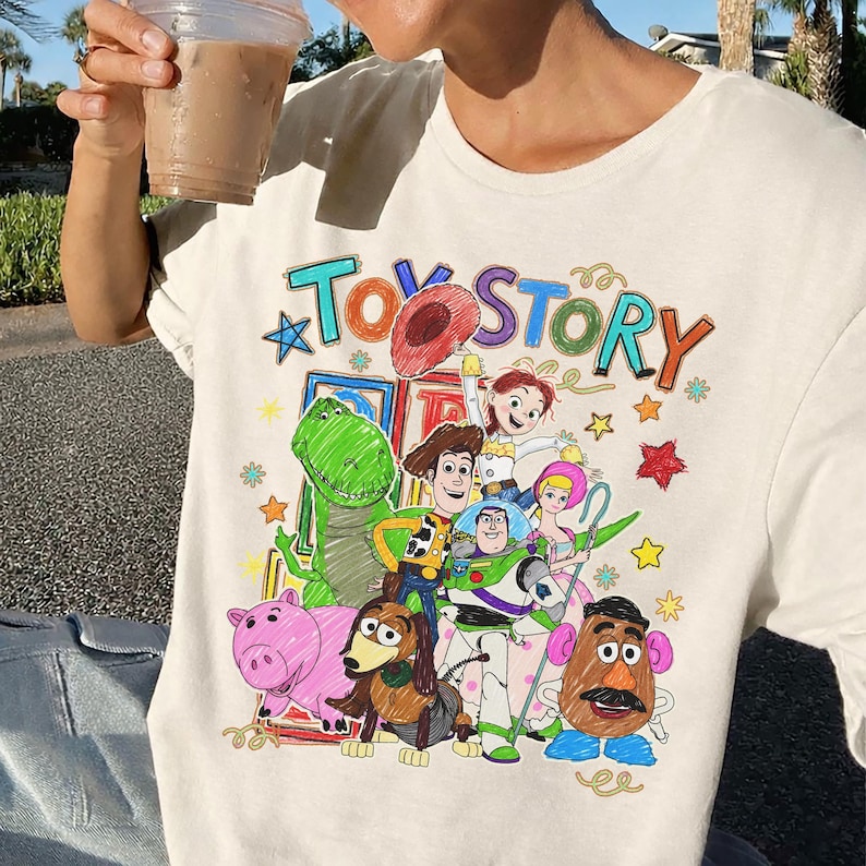 Może przedstawiać: Kremowy t-shirt z kolorową grafiką postaci z filmu animowanego "Toy Story". Projekt zawiera tytuł filmu i r&oacute;żne postacie, takie jak Woody, Buzz Lightyear i Rex. Koszulka to element odzieży codziennej.