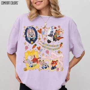 May include: A light purple Comfort Colors t-shirt featuring cartoon characters from Zootopia. The design includes Judy Hopps, Nick Wilde, and other characters in frames, a police vehicle, and the text "WELCOME TO ZOOOTOPIA".