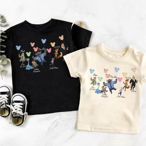 May include: A black t-shirt and a cream t-shirt, each featuring a graphic print of Zootopia characters holding colourful Mickey Mouse-shaped balloons. The characters include Flash, Chief Bogo, Nick Wilde, Judy Hopps, and others with their names printed below.