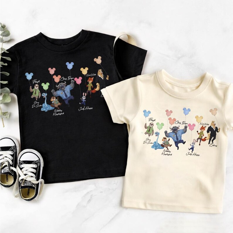 May include: A black t-shirt and a cream t-shirt, each featuring a graphic print of Zootopia characters holding colourful Mickey Mouse-shaped balloons. The characters include Flash, Chief Bogo, Nick Wilde, Judy Hopps, and others with their names printed below.