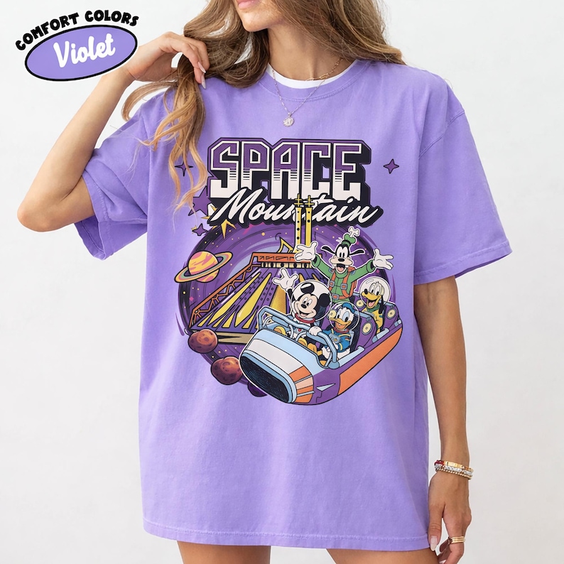 May include: Light purple Comfort Colors t-shirt with a Space Mountain graphic. The design features Mickey Mouse, Donald Duck, and Goofy on a ride, with a planet and stars. The words 'Space Mountain' are in white and purple.