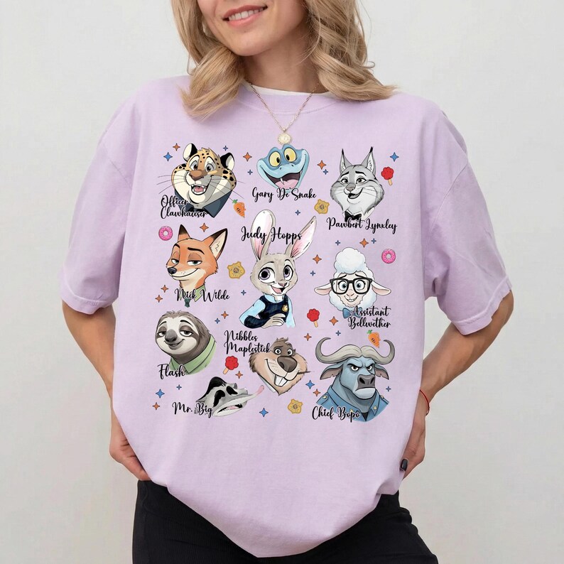 May include: A light purple short-sleeve t-shirt featuring a graphic print of various animated animal characters from Zootopia, including Judy Hopps, Nick Wilde, and Flash, with their names printed in black script text alongside small colorful icons.