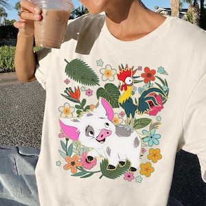 Puede incluir: Camiseta color crema con una ilustraci&oacute;n de dibujos animados de un cerdo blanco con orejas rosas y un gallo sobre su espalda, rodeado de flores y follaje coloridos. El dise&ntilde;o es vibrante y detallado.