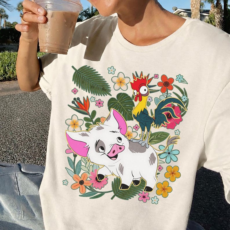 Puede incluir: Camiseta color crema con una ilustraci&oacute;n de dibujos animados de un cerdo blanco con orejas rosas y un gallo sobre su espalda, rodeado de flores y follaje coloridos. El dise&ntilde;o es vibrante y detallado.
