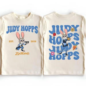 May include: A cream-colored t-shirt featuring Judy Hopps from Zootopia. The front displays the character with the text Judy Hopps, Est. 2016, and Zootopia. The back shows the character surrounded by repeating blue text and small icons.