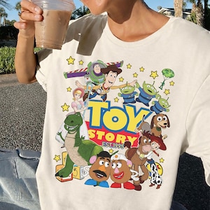 May include: Cream-colored t-shirt featuring a colorful graphic of Toy Story characters, including Woody, Buzz Lightyear, and the Little Green Men. The words "TOY STORY EST. 1995" are also displayed. The design is surrounded by yellow stars.