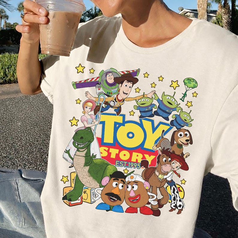 May include: Cream-colored t-shirt featuring a colorful graphic of Toy Story characters, including Woody, Buzz Lightyear, and the Little Green Men. The words "TOY STORY EST. 1995" are also displayed. The design is surrounded by yellow stars.