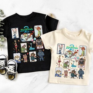 May include: Two short-sleeve t-shirts, one black and one cream, each featuring a graphic print of the Zootopia 2 logo surrounded by various character illustrations in individual frames, including Judy Hopps, Nick Wilde, and other animals.