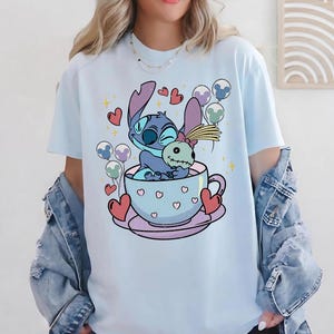 May include: A light blue t-shirt featuring a graphic of the character Stitch sitting in a light blue teacup while holding a small doll. The design includes red hearts, colorful balloons with Mickey Mouse ears, and yellow stars surrounding the teacup.