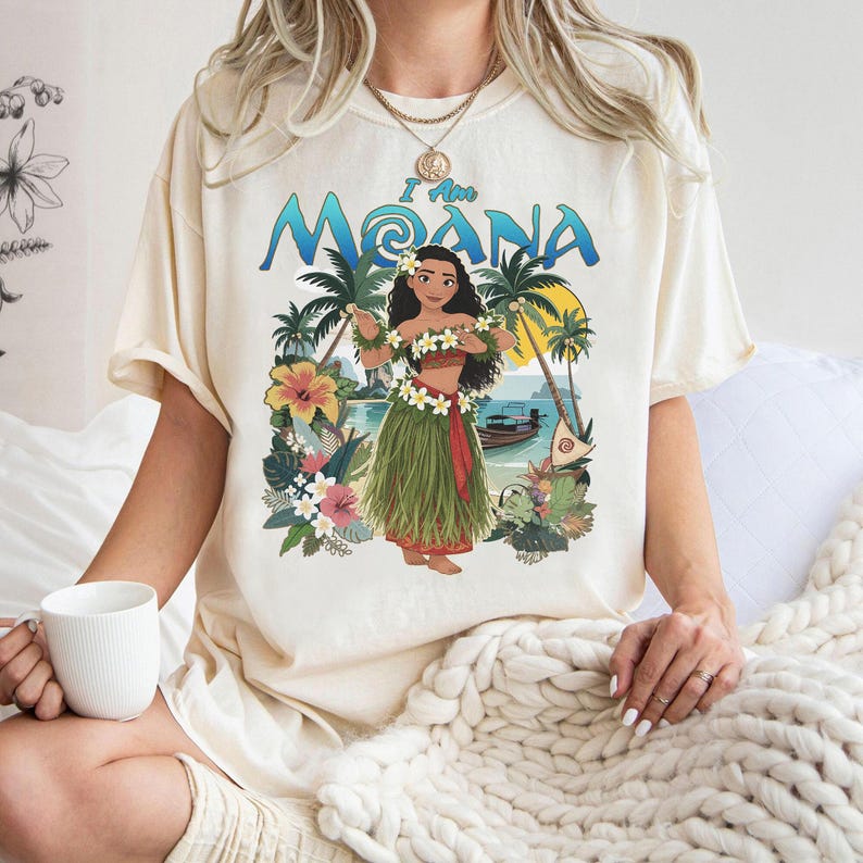 May include: Cream-colored t-shirt featuring a graphic of a woman in a grass skirt with the text "I Am Moana." The design includes palm trees, flowers, and a boat on a beach. The shirt has short sleeves and a relaxed fit.