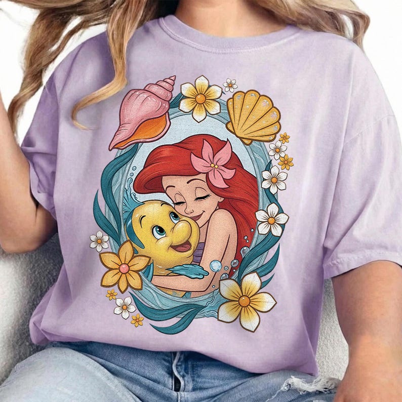 May include: A lilac-colored t-shirt featuring a graphic of Ariel from The Little Mermaid hugging Flounder. The design is framed by a blue oval with seashells and flowers. The shirt has short sleeves.